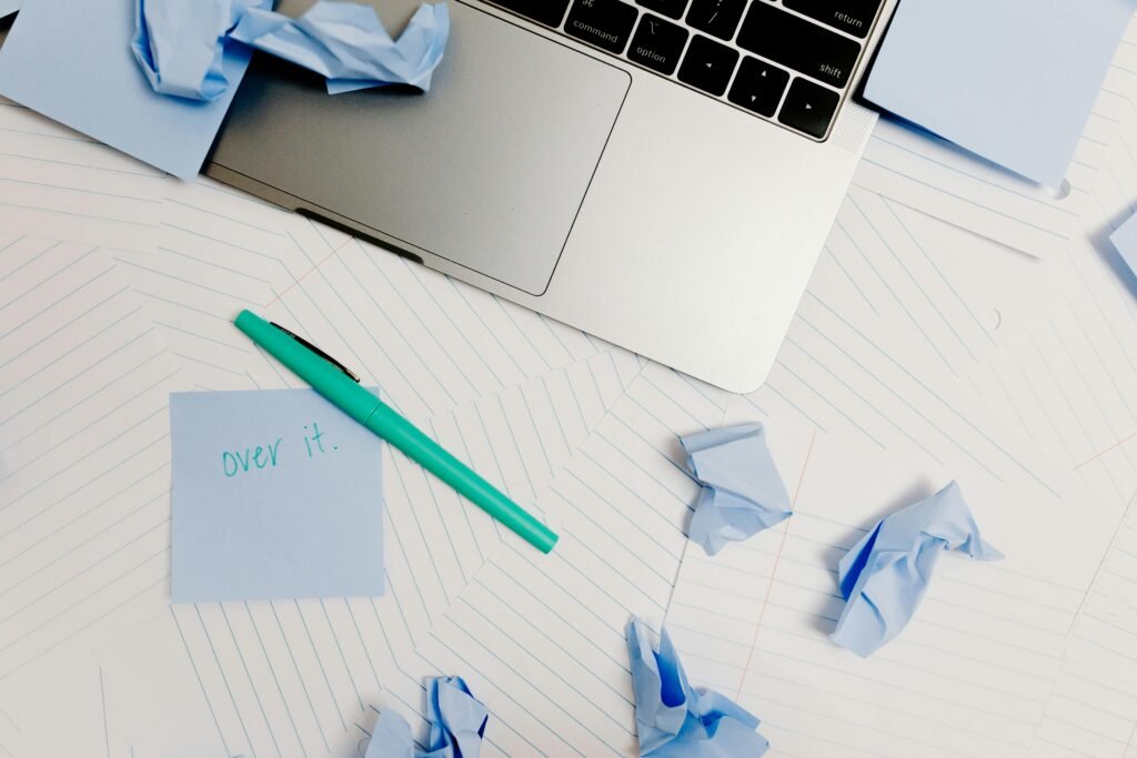 A messy desk with a laptop, post-it note saying 'over it,' and crumpled notes symbolizing burnout.