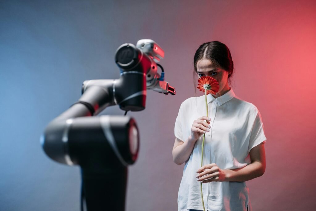 A woman smelling a red flower next to a robot arm, highlighting human-technology interaction.